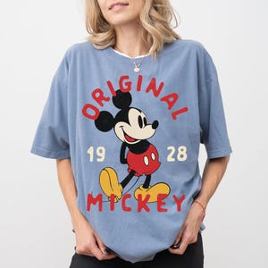 May include: A light blue t-shirt featuring a large graphic of Mickey Mouse. The design includes the word "ORIGINAL" in red, the number "1928", and the word "MICKEY" in red. Mickey Mouse is depicted in his classic outfit.