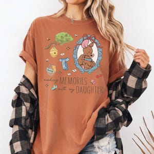 Op de afbeelding: Roestkleurig T-shirt met een cartoonillustratie van een beer en de tekst "making MEMORIES with my DAUGHTER". Het ontwerp bevat een honingpot, een bijenkorf en andere grillige elementen. Het shirt heeft opgerolde mouwen.
