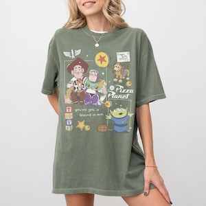 Pu&ograve; includere: T-shirt verde oliva con una grafica dei personaggi di Toy Story, tra cui Woody, Buzz Lightyear e i piccoli omini verdi. La maglietta presenta anche il testo "Pizza Planet" e "You've got a friend in me."