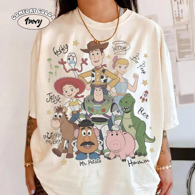 May include: An ivory t-shirt featuring a graphic print of Toy Story characters including Woody, Buzz Lightyear, Jessie, Bo Peep, Forky, Bullseye, Mr. Potato Head, Rex, and Hamm, with their names written in cursive script and small yellow stars.