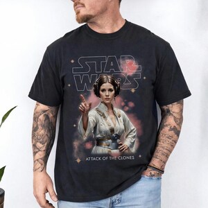 Peut inclure: T-shirt noir avec un graphisme d'une femme en tenue blanche, le texte "STAR WARS" et "ATTACK OF THE CLONES". Le design a un aspect vintage et vieilli.