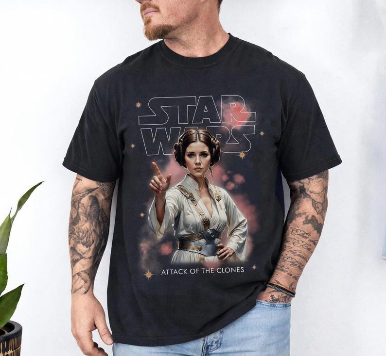 Peut inclure: T-shirt noir avec un graphisme d'une femme en tenue blanche, le texte "STAR WARS" et "ATTACK OF THE CLONES". Le design a un aspect vintage et vieilli.
