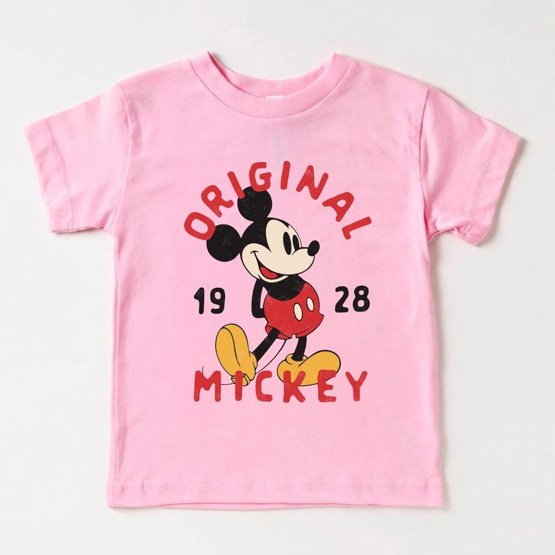 May include: Pink t-shirt featuring Mickey Mouse with the text "ORIGINAL MICKEY 1928" in red. Mickey Mouse is depicted in his classic pose, wearing red shorts, yellow shoes, and black gloves.