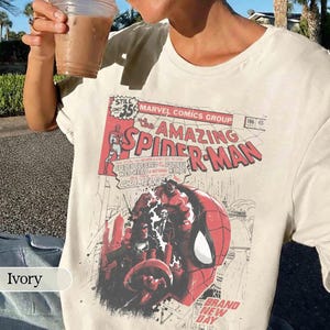 Może przedstawiać: Kremowa koszulka z nadrukiem z okładki komiksu "The Amazing Spider-Man". Grafika zawiera twarz Spider-Mana i napis "BRAND NEW DAY". Koszulka ma okrągły dekolt i kr&oacute;tkie rękawy.