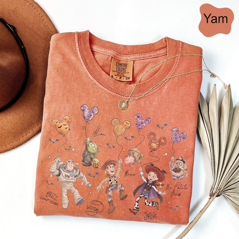 Puede incluir: Una camiseta de cuello redondo Comfort Colors color &ntilde;ame con un gr&aacute;fico de personajes de Toy Story sosteniendo globos con forma de Mickey Mouse. Los personajes incluyen a Buzz Lightyear, Woody, Jessie y Mr. Potato Head. Un collar de oro est&aacute; drapeado sobre la camisa.