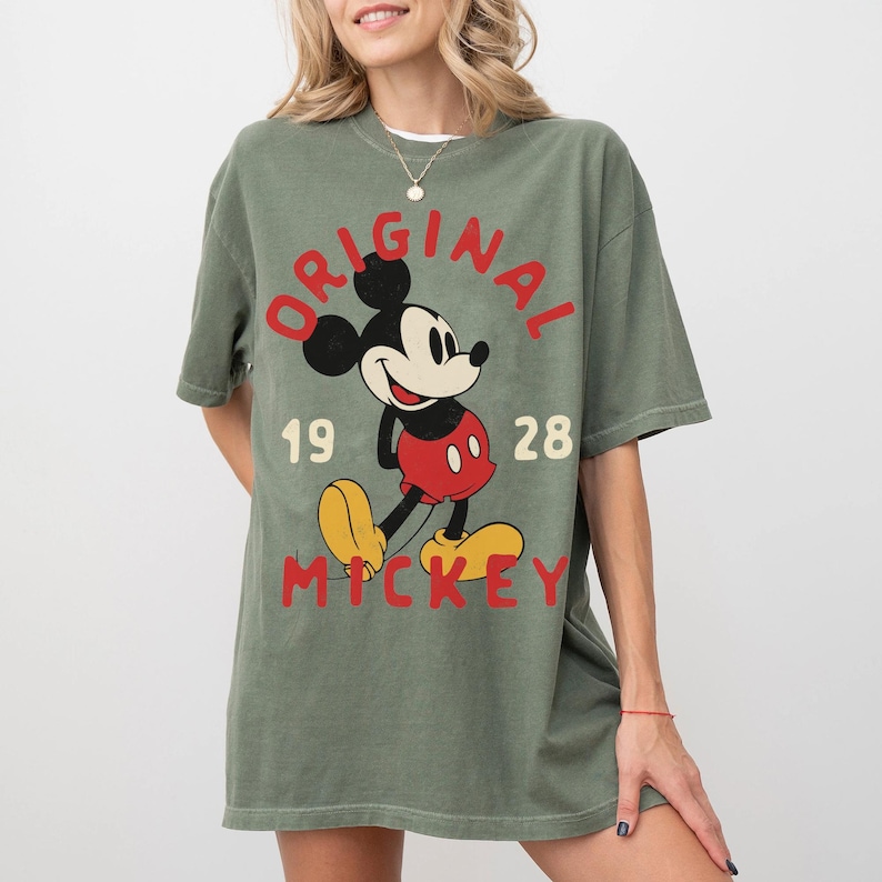 May include: Olive green t-shirt featuring a graphic of Mickey Mouse with the text "ORIGINAL MICKEY 1928". The cartoon character is in red, black, yellow, and white. The shirt has short sleeves and a relaxed fit.