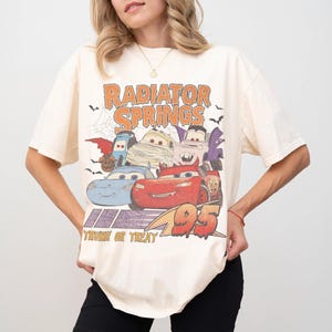 May include: Cream-colored t-shirt featuring a graphic of the Radiator Springs characters in Halloween costumes. The text "Radiator Springs" is above the cars, and "Trunk or Treat" is below. The number "95" is also visible.