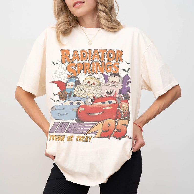 May include: Cream-colored t-shirt featuring a graphic of the Radiator Springs characters in Halloween costumes. The text "Radiator Springs" is above the cars, and "Trunk or Treat" is below. The number "95" is also visible.
