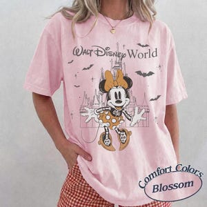 May include: Pale pink t-shirt featuring a Halloween-themed Minnie Mouse graphic with a castle backdrop. The text "Walt Disney World" is above the graphic, and "Comfort Colors Blossom" is printed at the bottom.