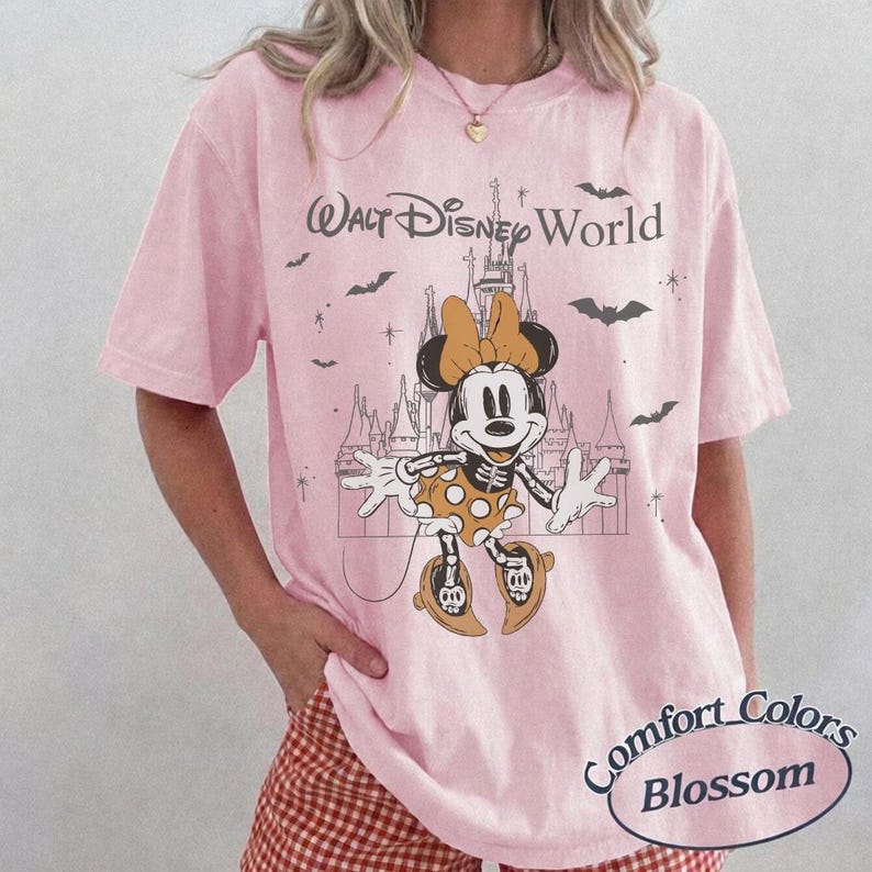 May include: Pale pink t-shirt featuring a Halloween-themed Minnie Mouse graphic with a castle backdrop. The text "Walt Disney World" is above the graphic, and "Comfort Colors Blossom" is printed at the bottom.