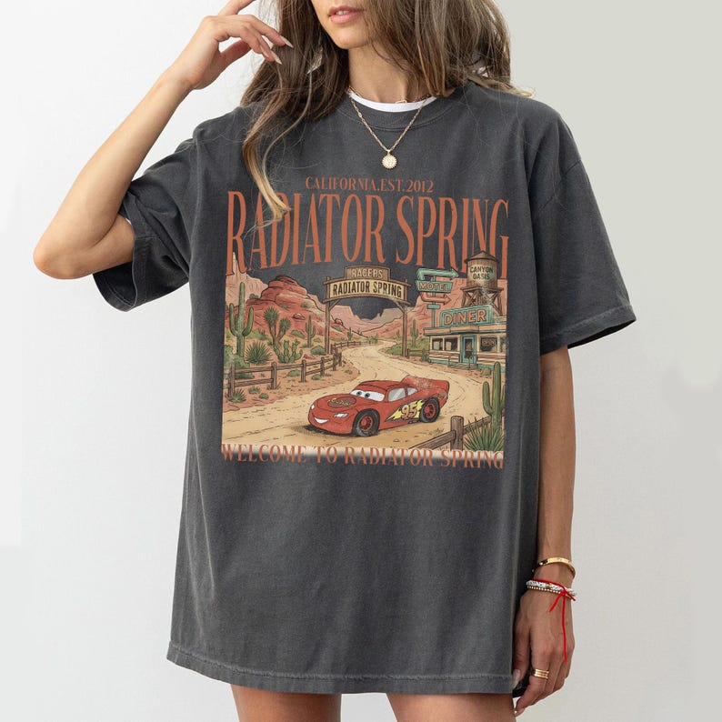 May include: Dark gray t-shirt featuring a graphic of Radiator Springs from the movie Cars. The design includes a red race car, desert scenery, and the text "CALIFORNIA EST. 2012 RADIATOR SPRING WELCOME TO RADIATOR SPRING."