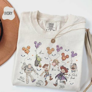 Puede incluir: Camiseta Comfort Colors de color marfil con un gr&aacute;fico de personajes de Disney sosteniendo globos con forma de Mickey Mouse. Los personajes incluyen a Buzz Lightyear, Woody y Jessie. La camiseta tiene cuello redondo y mangas cortas.