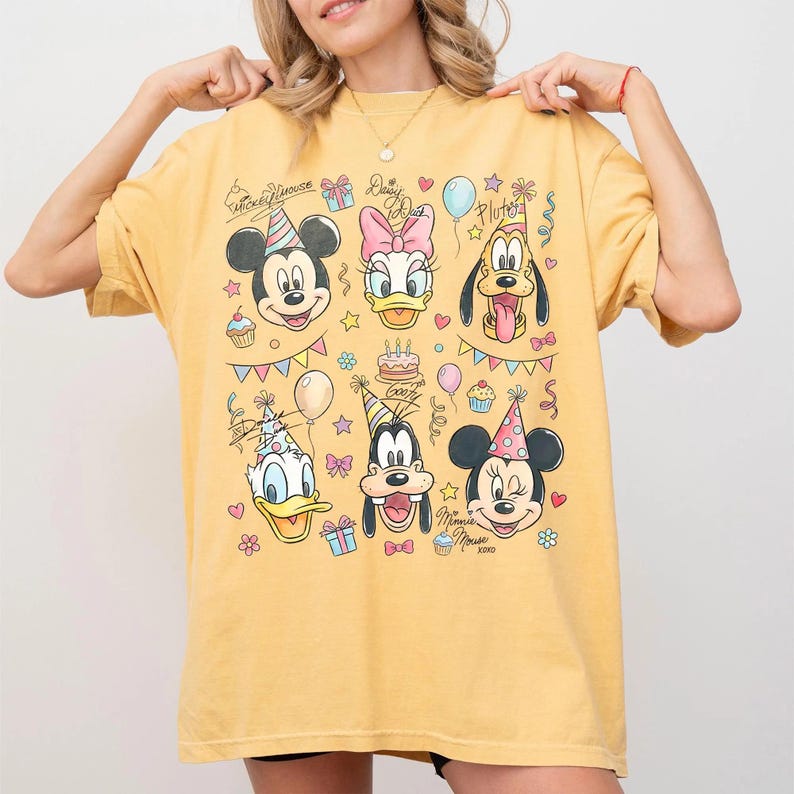 Puede incluir: Camiseta amarilla mostaza con un dise&ntilde;o de Mickey Mouse, Daisy Duck, Pluto, Donald Duck, Goofy y Minnie Mouse. El dise&ntilde;o incluye gorros de fiesta, globos, cupcakes y el texto "Mickey Mouse" y "Daisy Duck".