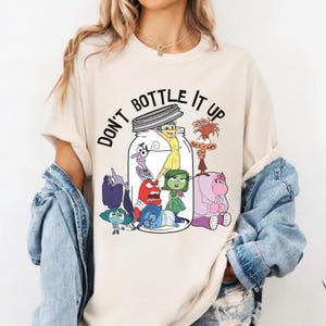 Peut inclure: Un t-shirt crème avec l'inscription "DON'T BOTTLE IT UP" au-dessus d'un dessin animé de personnages à l'intérieur d'un bocal. Les personnages sont de différentes couleurs et poses. Le t-shirt a des manches courtes et un col rond.