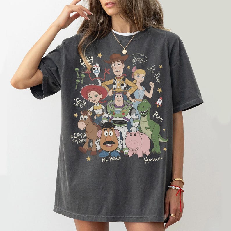 May include: A charcoal gray short-sleeve t-shirt featuring a graphic print of Toy Story characters including Woody, Buzz Lightyear, Jessie, Bo Peep, Bullseye, Mr. Potato Head, Hamm, Rex, and Forky, with their names written in a cursive script.