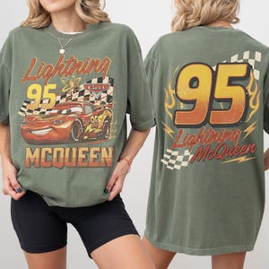 May include: An olive green t-shirt featuring Lightning McQueen from the Cars film. The front graphic shows McQueen with the number 95 and the text "Lightning McQueen". The back of the shirt has the number 95 and "Lightning McQueen" with a checkered flag.