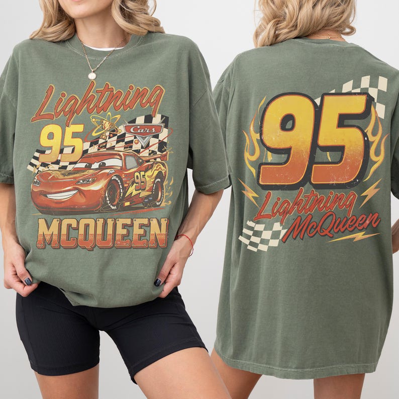 May include: An olive green t-shirt featuring Lightning McQueen from the Cars film. The front graphic shows McQueen with the number 95 and the text "Lightning McQueen". The back of the shirt has the number 95 and "Lightning McQueen" with a checkered flag.