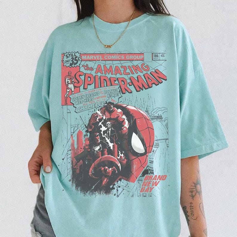 Może przedstawiać: Jasnoniebieska koszulka z komiksową ilustracją Spider-Mana. Projekt zawiera tekst "the AMAZING SPIDER-MAN" i "MARVEL COMICS GROUP". Grafika jest w kolorze czerwonym, czarnym i białym.