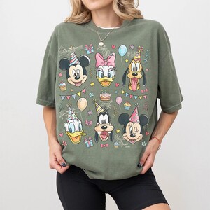 Puede incluir: Camiseta verde oliva con un gr&aacute;fico colorido de Mickey Mouse, Daisy Duck, Pluto, Donald Duck, Goofy y Minnie Mouse, todos con sombreros de fiesta. El dise&ntilde;o incluye globos, confeti y el texto "Mickey Mouse" y "Minnie Mouse".