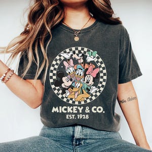 Puede incluir: Camiseta gris oscuro con un dise&ntilde;o circular de personajes de Disney, incluyendo Mickey y Minnie Mouse, Donald Duck y Goofy. El dise&ntilde;o est&aacute; rodeado por un patr&oacute;n de cuadros blancos y negros. El texto "MICKEY & CO. EST. 1928" est&aacute; impreso debajo.