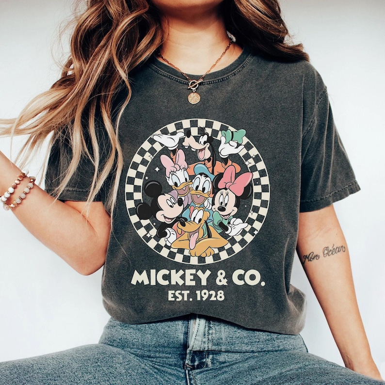 Puede incluir: Camiseta gris oscuro con un dise&ntilde;o circular de personajes de Disney, incluyendo Mickey y Minnie Mouse, Donald Duck y Goofy. El dise&ntilde;o est&aacute; rodeado por un patr&oacute;n de cuadros blancos y negros. El texto "MICKEY & CO. EST. 1928" est&aacute; impreso debajo.