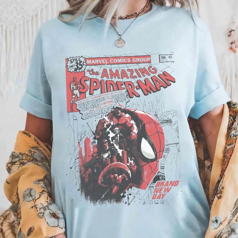 Może przedstawiać: Jasnoniebieska koszulka z okładką komiksu The Amazing Spider-Man w stylu vintage. Grafika zawiera logo Spider-Mana, tekst komiksu oraz czerwoną i czarną ilustrację twarzy Spider-Mana i innych postaci. Rękawy są podwinięte.