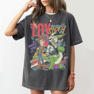 May include: Dark grey t-shirt with the Toy Story logo and characters Woody, Buzz Lightyear, and Rex. The design includes the text "TOY STORY" and "SINCE 1995" in a colourful, retro style.