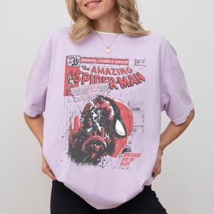 Może przedstawiać: Jasnofioletowa koszulka z okładką komiksu The Amazing Spider-Man w stylu vintage. Grafika zawiera tytuł komiksu, twarz Spider-Mana i inne elementy komiksu. Koszulka ma kr&oacute;tkie rękawy.