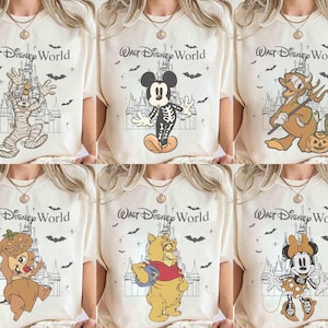 May include: Cream-coloured t-shirts featuring Halloween-themed Disney characters. Designs include a mummy Goofy, skeleton Mickey Mouse, devilish Donald Duck, Chip with a pumpkin, Winnie the Pooh, and Minnie Mouse. Each shirt has the text "Walt Disney World".
