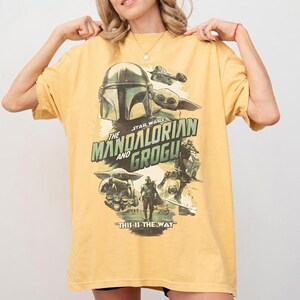 K&ouml;nnte beinhalten: Senfgelbes T-Shirt mit einer Grafik aus der Star Wars-Serie "The Mandalorian and Grogu". Das Design enth&auml;lt Bilder des Mandalorianer-Helms, Grogu und anderer Charaktere mit dem Satz "This is the Way."