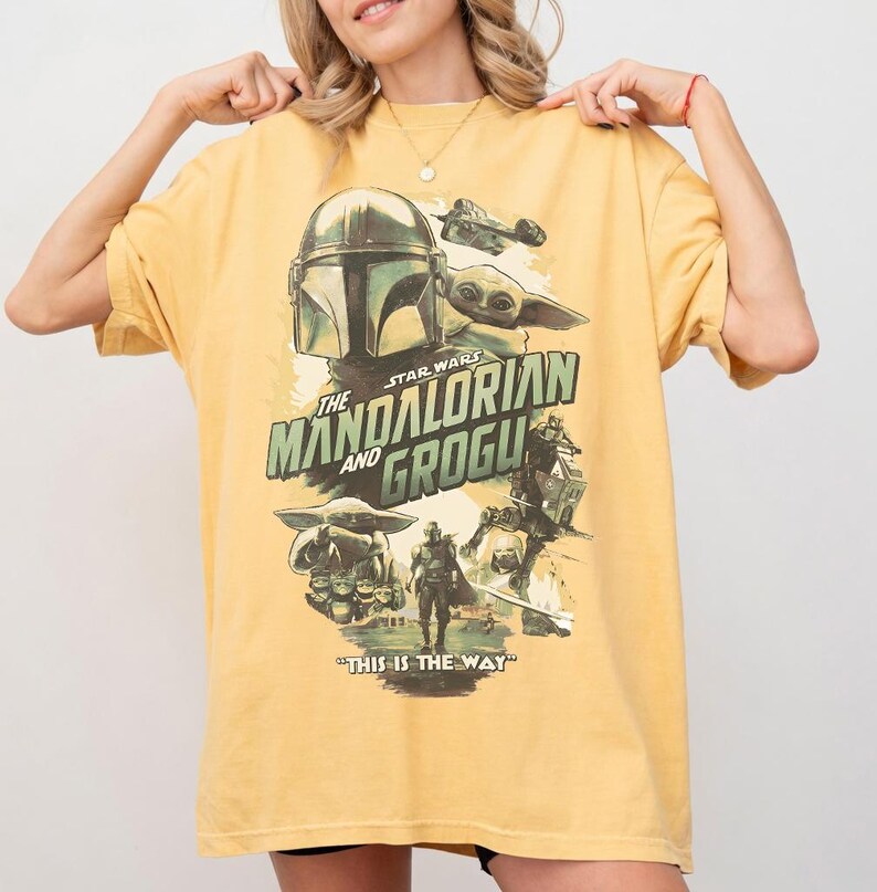 K&ouml;nnte beinhalten: Senfgelbes T-Shirt mit einer Grafik aus der Star Wars-Serie "The Mandalorian and Grogu". Das Design enth&auml;lt Bilder des Mandalorianer-Helms, Grogu und anderer Charaktere mit dem Satz "This is the Way."