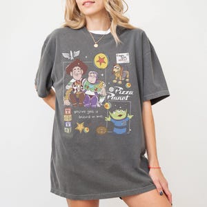 Pu&ograve; includere: T-shirt grigio scuro con una grafica dei personaggi del film Toy Story, tra cui Woody, Buzz Lightyear e gli omini verdi. La maglietta presenta anche il testo "Pizza Planet" e "You've got a friend in me."