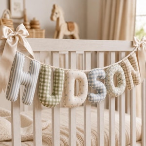 May include: A decorative fabric banner spelling the name HUDSON, featuring stuffed letters in various patterns including blue stripes, green gingham, light blue stars, blue gingham, floral print, and tan plaid, tied with beige satin ribbons at each end.