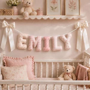 May include: A decorative fabric banner spelling the name EMILY in stuffed, plush letters. Each letter features a different pink and white pattern, including gingham, polka dots, and floral prints, suspended by a white rope with large satin bows.