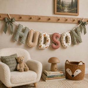 May include: A fabric letter banner spelling HUDSON in various patterns including green gingham, beige stripes, fox prints, red gingham, and pine tree motifs. The banner is hung on a wooden peg rail with olive green ribbon bows at each end.