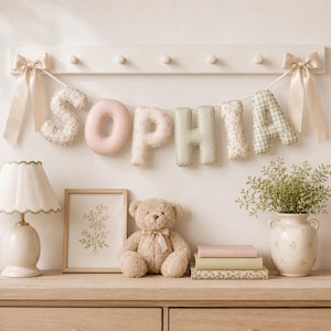 May include: A fabric letter banner spelling the name SOPHIA, featuring soft pastel colors and floral, gingham, and striped patterns. The letters are strung on a cord with beige satin bows at each end, suspended from a white wooden wall rack.