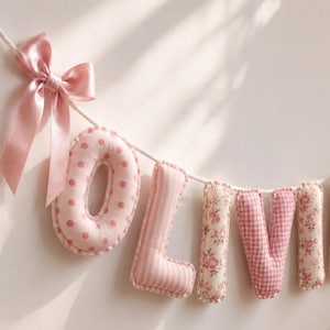 May include: A decorative fabric banner spelling the name OLIVIA with stuffed, plush letters hanging from a white rope. The letters feature various pink and white patterns including polka dots, vertical stripes, floral prints, and gingham, finished with a pink satin bow.
