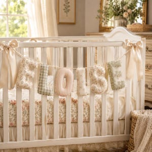 May include: A decorative fabric banner spelling the name ELOISE, featuring plush, stuffed letters in alternating patterns of floral prints, gingham checks, and stripes in soft pastel tones, suspended by a rope with large cream satin bows at each end.