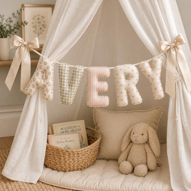 May include: A decorative fabric banner spelling the name AVERY with individual stuffed letters in various patterns including floral, gingham, stripes, hearts, and bows, suspended by a rope with beige satin bows on each end of the display.