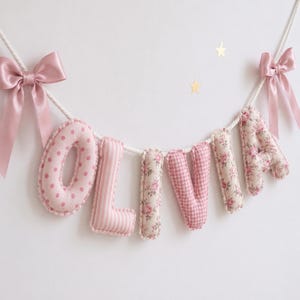 May include: A decorative fabric banner spelling the name OLIVIA with stuffed, padded letters suspended on a white rope. Each letter features a different pink pattern, including polka dots, stripes, floral prints, and gingham, finished with pink satin bows.
