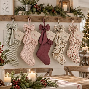 Op de afbeelding: Vijf stoffen kerstsokken hangend aan een houten schoorsteenmantel, elk voorzien van een decoratieve strik en een gepersonaliseerd naamplaatje. De sokken hebben diverse patronen, waaronder groene strepen, rode stippen, effen rood, herten en bloemen.