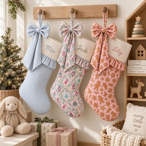 May include: Three Christmas stockings hanging from a wooden rack. The first is blue and white striped with the name Mia. The second features a floral pattern with the name Jack. The third has a gingerbread print with the name Lily. Each has a bow.