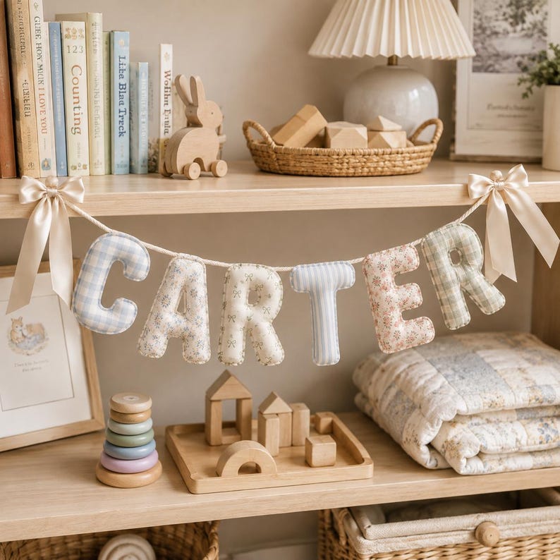 May include: A fabric letter banner spelling the name CARTER, featuring stuffed letters in various pastel patterns including blue gingham, floral prints, and light blue stripes, suspended by a white cord with cream-colored satin bows at each end.