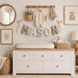 May include: A white wooden dresser with five drawers and a changing pad on top. Above, a banner spells MASON with fabric letters in green, beige, and blue patterns. A beige baby romper hangs on a wooden rack with a small basket and a cloth.