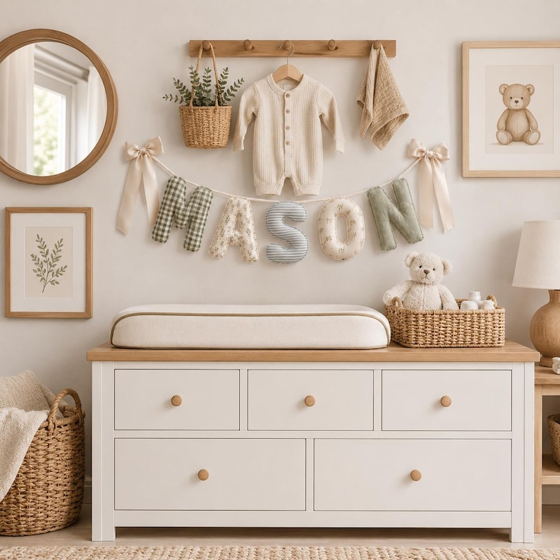 May include: A white wooden dresser with five drawers and a changing pad on top. Above, a banner spells MASON with fabric letters in green, beige, and blue patterns. A beige baby romper hangs on a wooden rack with a small basket and a cloth.