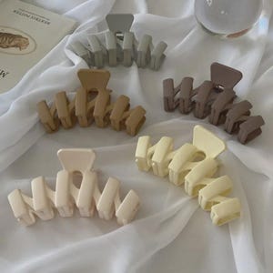 May include: A collection of five hair claw clips in various neutral colors: beige, light brown, taupe, gray, and pale yellow. The clips are made of matte plastic and have a zig-zag design. They are arranged on a white, textured surface.