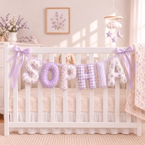 Pode incluir: Um berço branco apresenta uma guirlanda de tecido que soletra o nome SOPHIA em letras acolchoadas. As letras alternam entre padrões de cor lavanda, florais, de bolinhas e listrados, fixadas com fitas de cetim roxo claro nas extremidades.