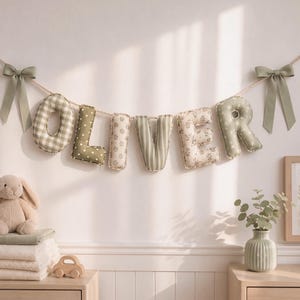 May include: A decorative banner spelling the name OLIVER with stuffed fabric letters in shades of sage green and cream. Each letter features a different pattern including gingham, polka dots, and floral prints, suspended by a rope with bows.