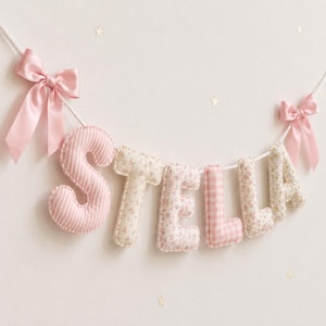 Puede incluir: Una guirnalda decorativa que deletrea el nombre STELLA con letras acolchadas cubiertas de tela en varios tonos de rosa, blanco y estampados florales. Las letras están ensartadas en un cordón blanco con un lazo de cinta de satén rosa en cada extremo.