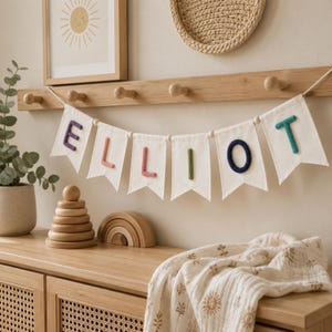 May include: A fabric banner featuring six white pennants hanging from a wooden peg rail. Each pennant displays a single letter in a different color, spelling out the name ELLIOT. The letters are purple, pink, coral, green, navy, and teal.